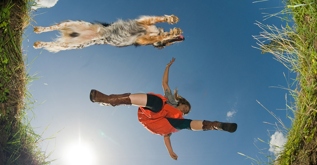 A dynamic view from below of a young woman jumps over a ditch with her sheepdog. 
